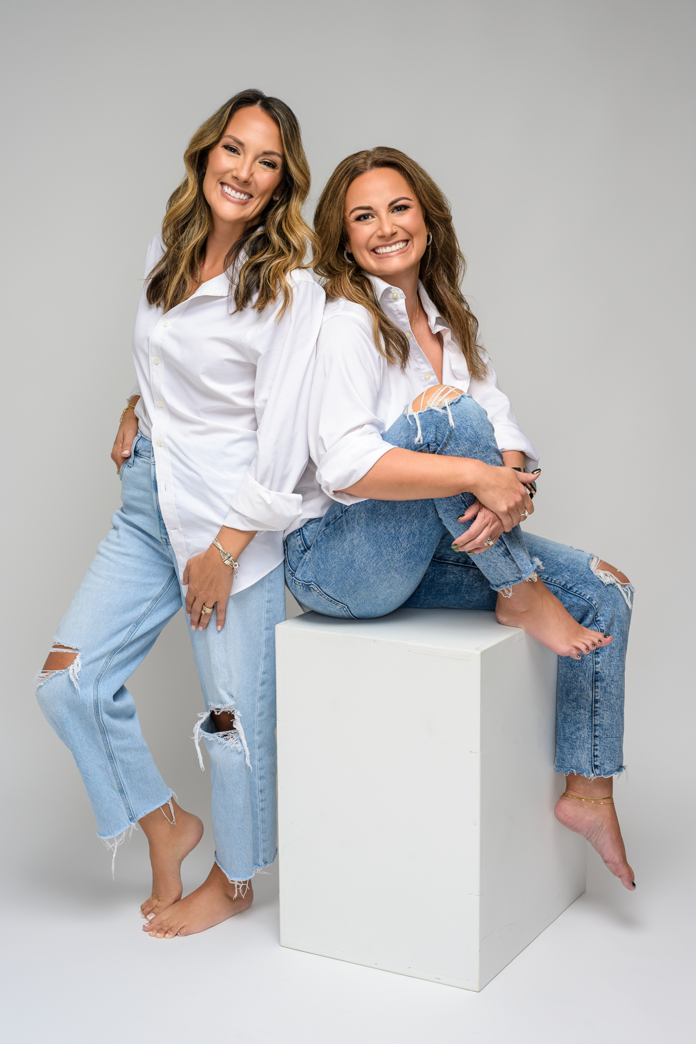 Stacey Matthys and Kristy Ruminski of The HOME Team posing barefoot in white shirts and ripped jeans against a light backdrop.