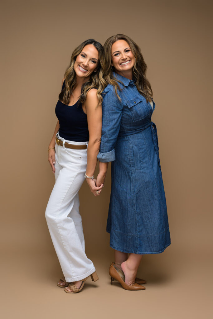 Stacey and Kristy seated back-to-back wearing fall-inspired outfits on a neutral brown background.