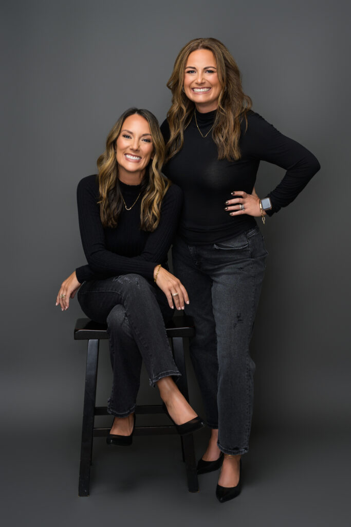 Kristy standing while Stacey sits on a black stool, both dressed in matching black turtlenecks and jeans.