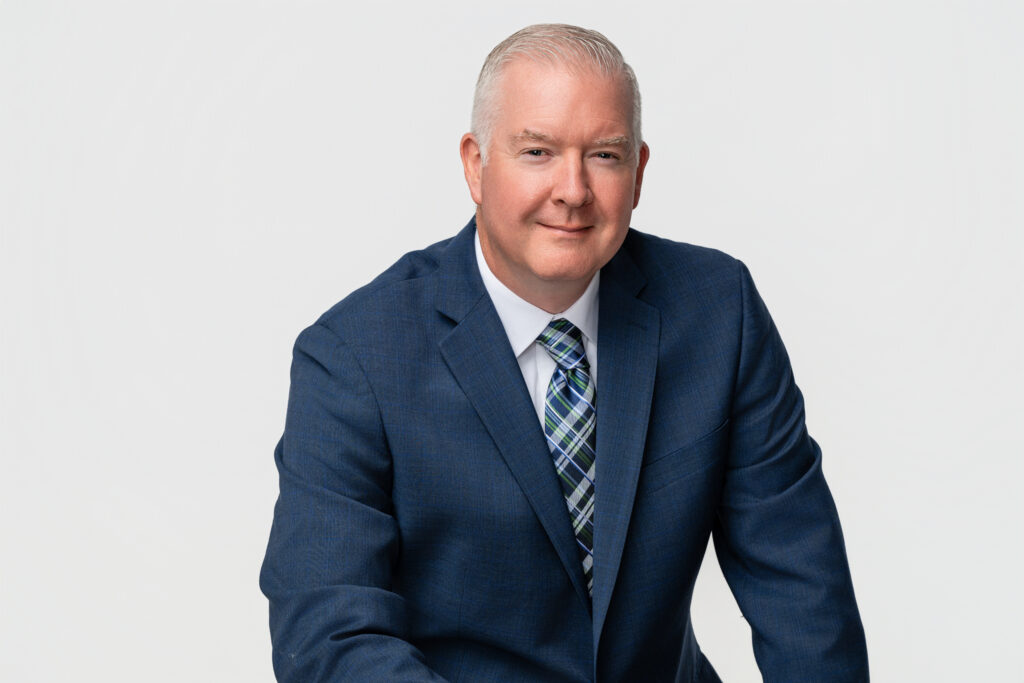 Professional headshot of a confident middle aged businessman in a navy suit and plaid tie, photographed in a South Bend studio.