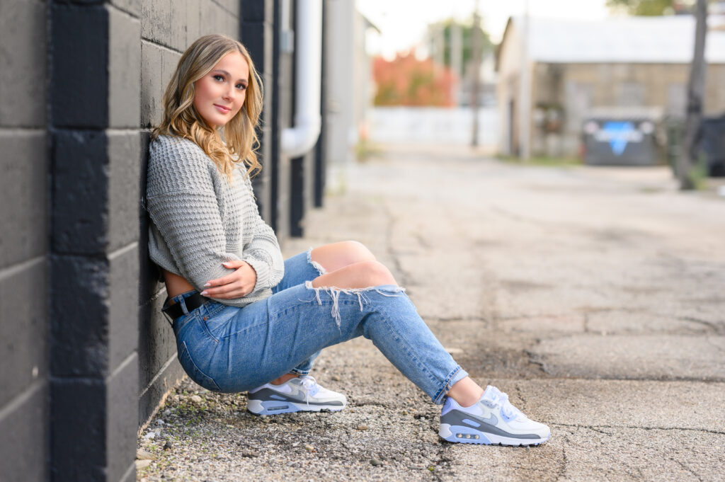 High school senior girl leaning against a brick wall in downtown Three Oaks Michigan during a modern senior portrait session, wearing ripped jeans and white Nikes.