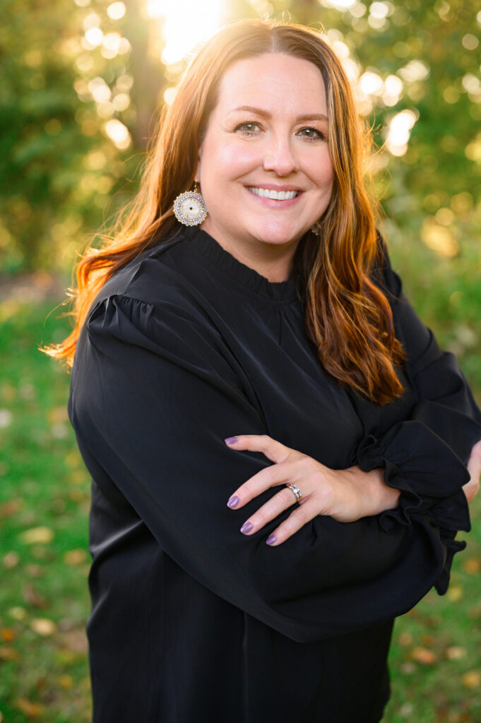Professional headshot of a smiling woman with long auburn hair, wearing a black blouse and statement earrings, taken outdoors near South Bend, Indiana.